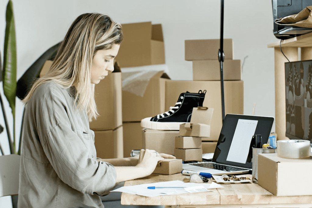 7 Essential Tips for Setting Up a WooCommerce for Service Business 4 A woman working at a desk with boxes and using a laptop for WooCommerce.